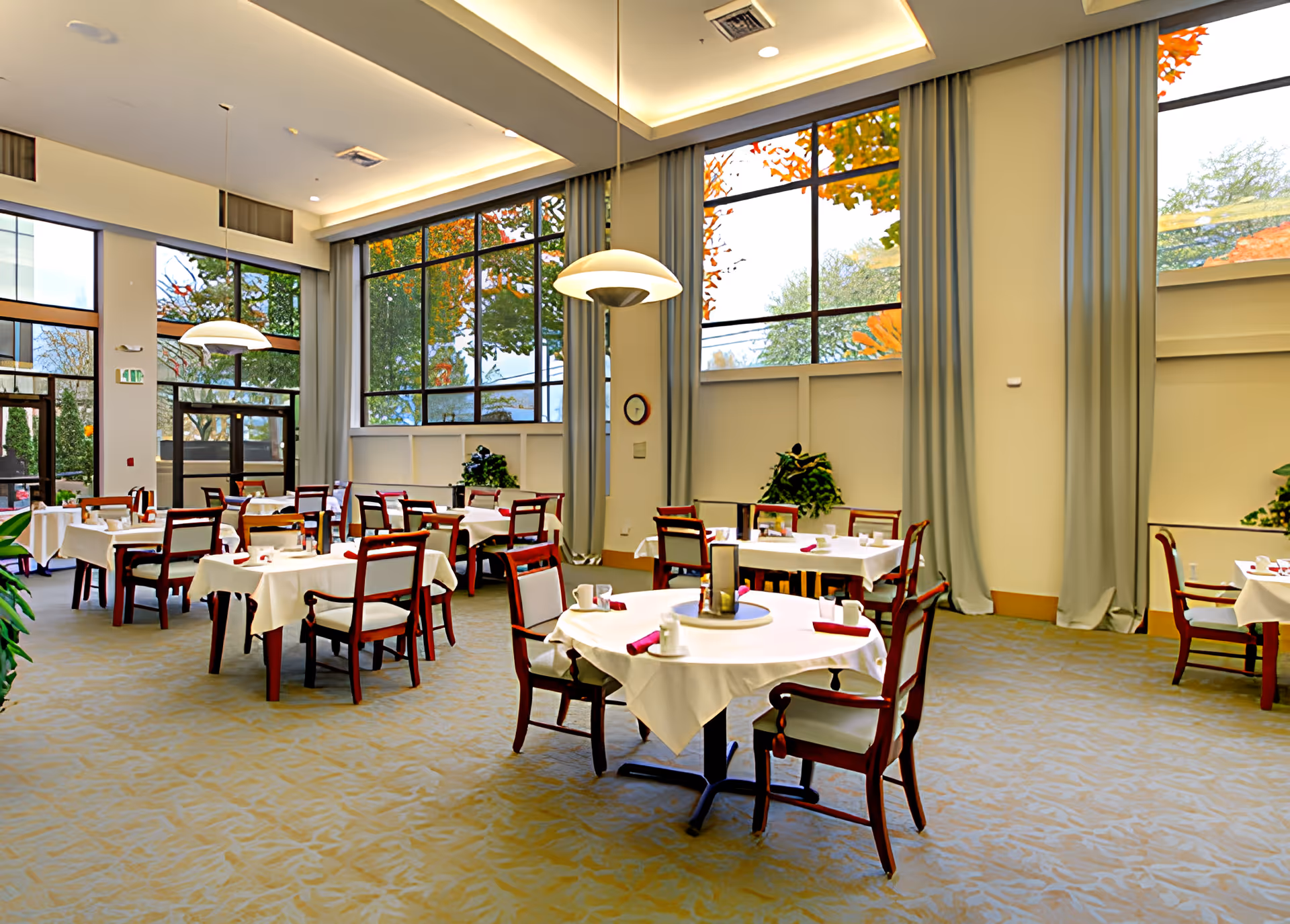 A bright dining room with multiple tables covered in white tablecloths, each set with cups, napkins, and condiments. The room features large windows with views of autumn trees outside, tall light blue curtains, and modern pendant lights hanging from the ceiling. The floor is carpeted in a light pattern, and there are some green plants placed around the room.