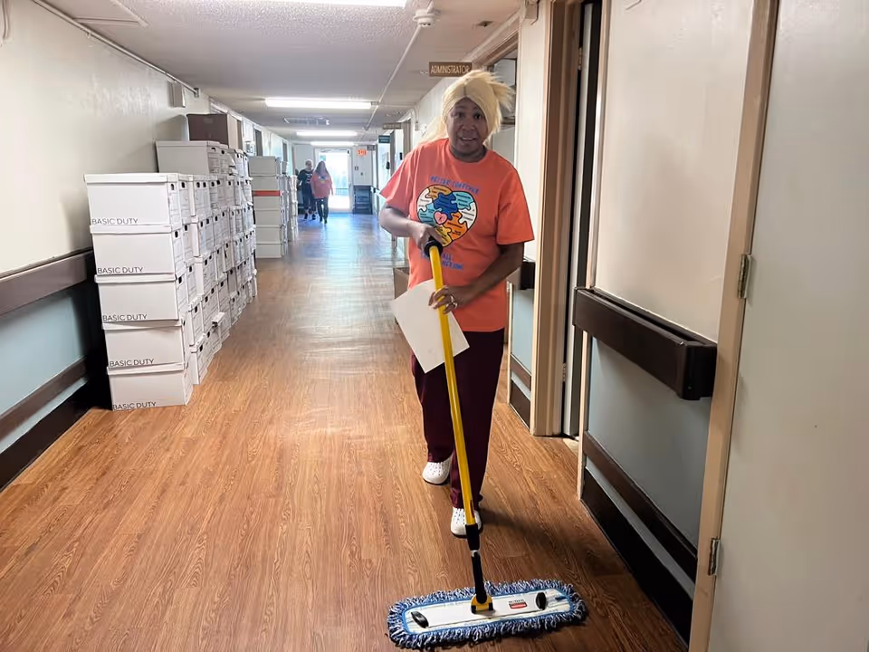 A woman with a yellow head wrap and an orange t-shirt is mopping the floor in a long hallway of a healthcare facility. The hallway has wooden flooring, beige walls, and several stacked boxes labeled 'BASIC DUTY' on the left side. There are doors along the right side of the hallway and a sign above indicating 'ADMINISTRATION'. Two other people are visible in the background walking down the hallway.