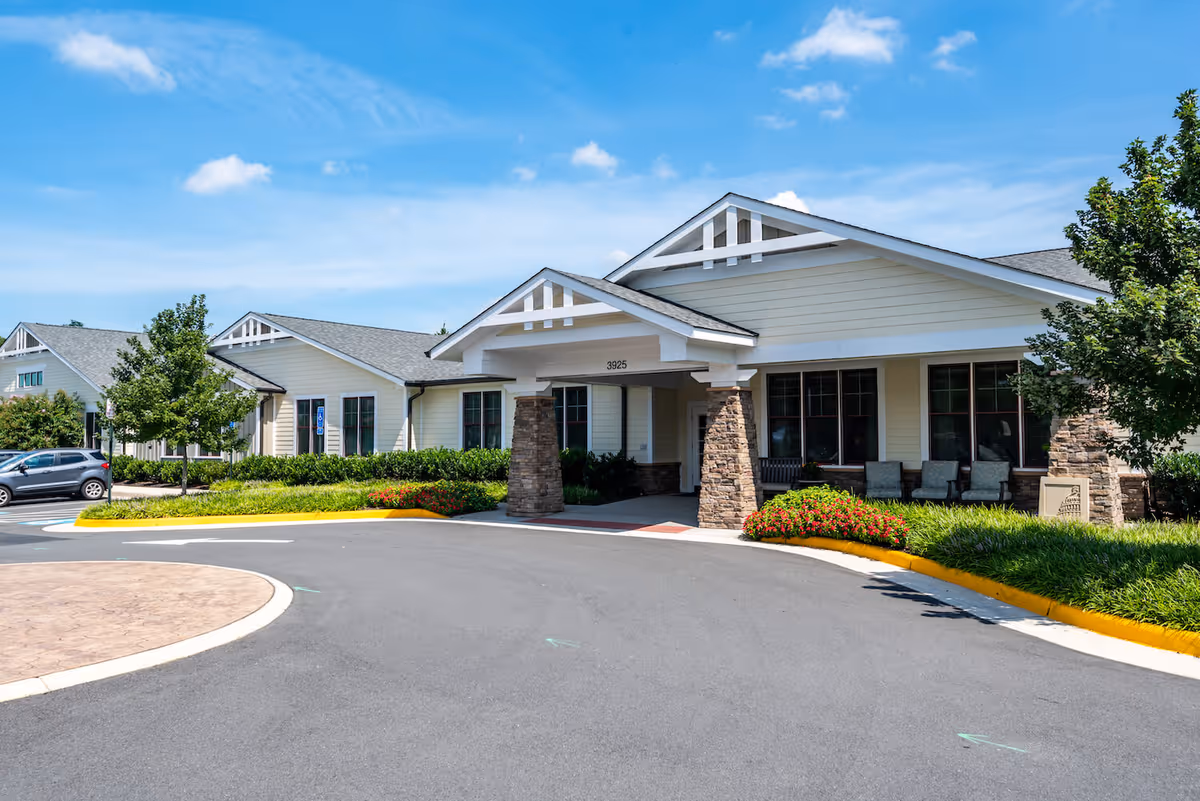 Front entrance of a single-story senior living facility with a covered portico, stone columns, landscaped flower beds, and a curved driveway.