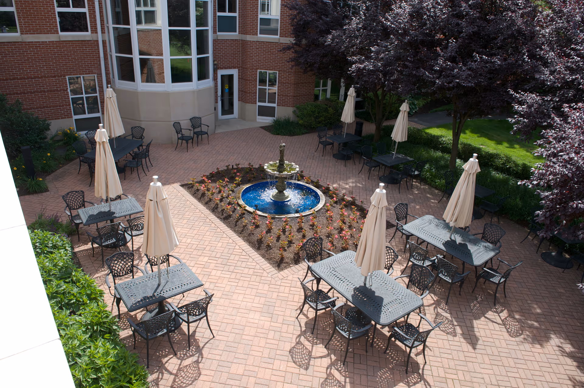 Outdoor courtyard area with several black metal tables and chairs, each table shaded by a closed beige umbrella. In the center is a circular water fountain surrounded by a flower bed. The courtyard is paved with brick and bordered by greenery and trees with dark purple leaves. A brick building with large windows is visible in the background.