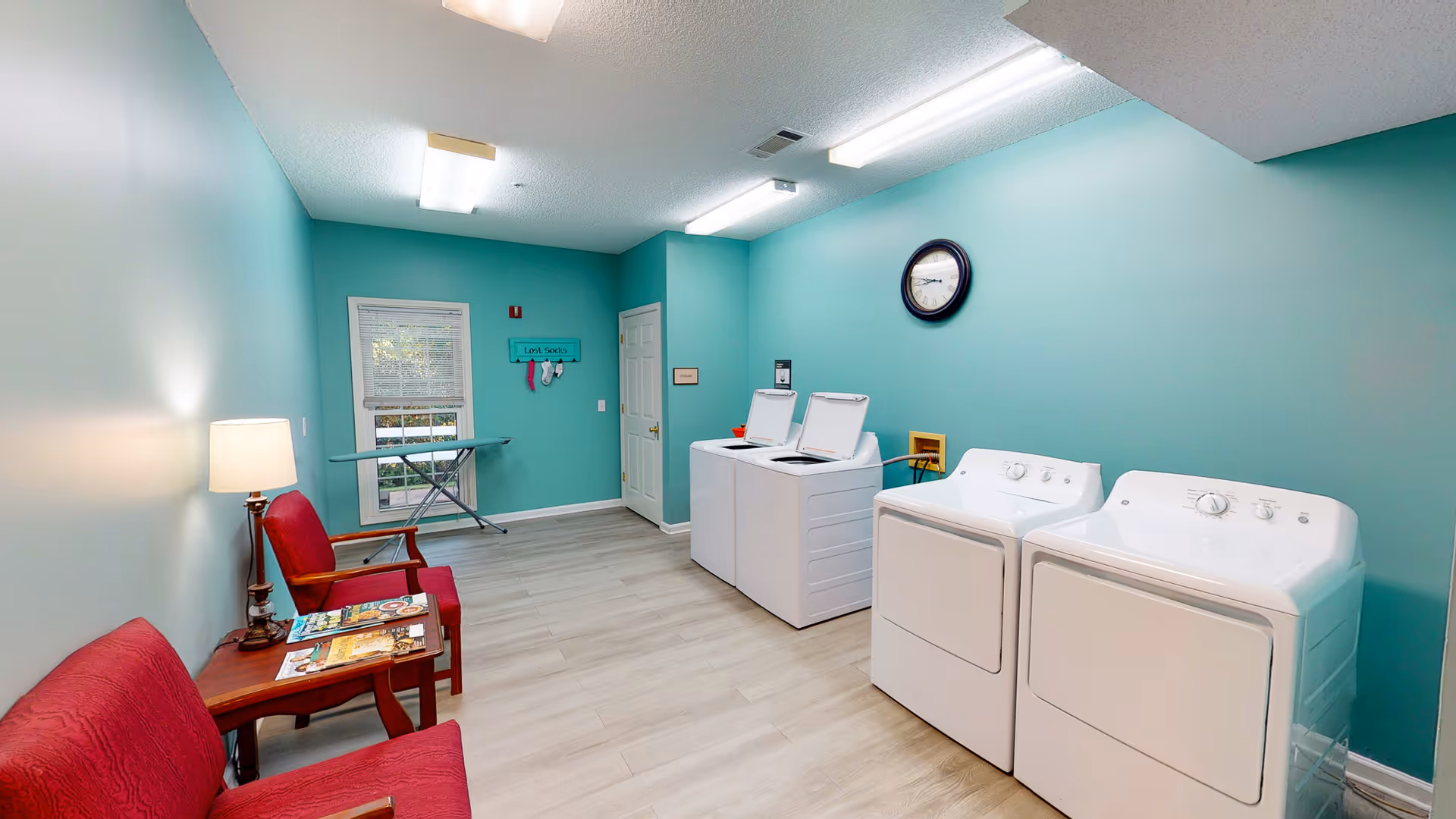 Laundry room with teal walls featuring two white top-loading washing machines and two white dryers. There are two red cushioned chairs with wooden arms and a small wooden table with magazines and a lamp. An ironing board is set up near a window with blinds. A clock hangs on the wall above the machines, and a sign labeled 'Lost Socks' with socks hanging below it is on the far wall.