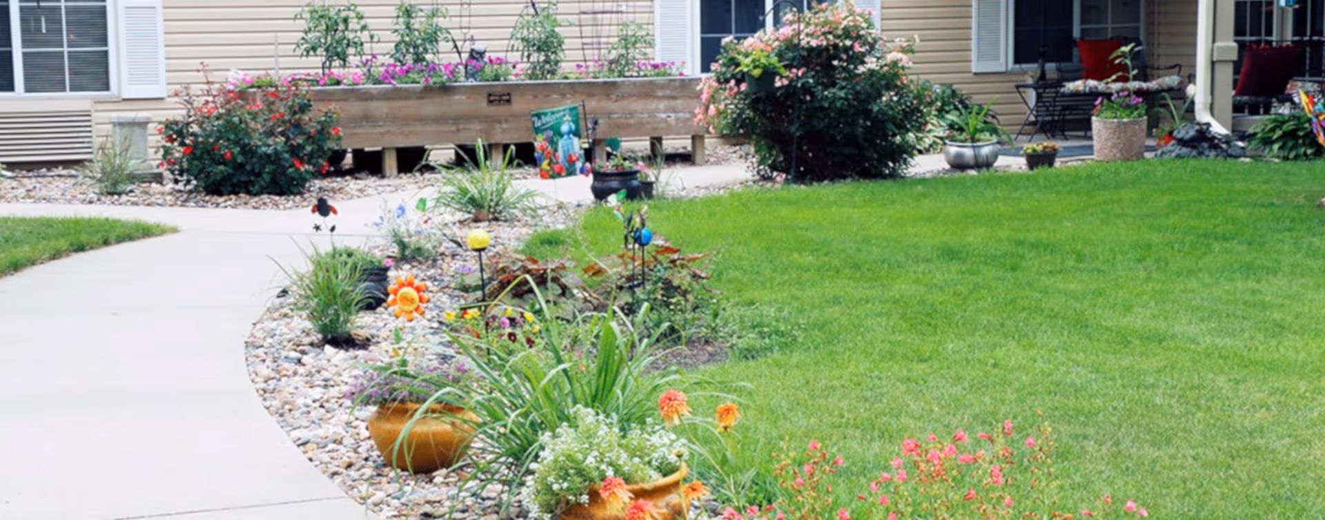 Curved concrete walkway bordered by rock gardens, potted flowers, and a green lawn leading to the front of a residential building with a porch.