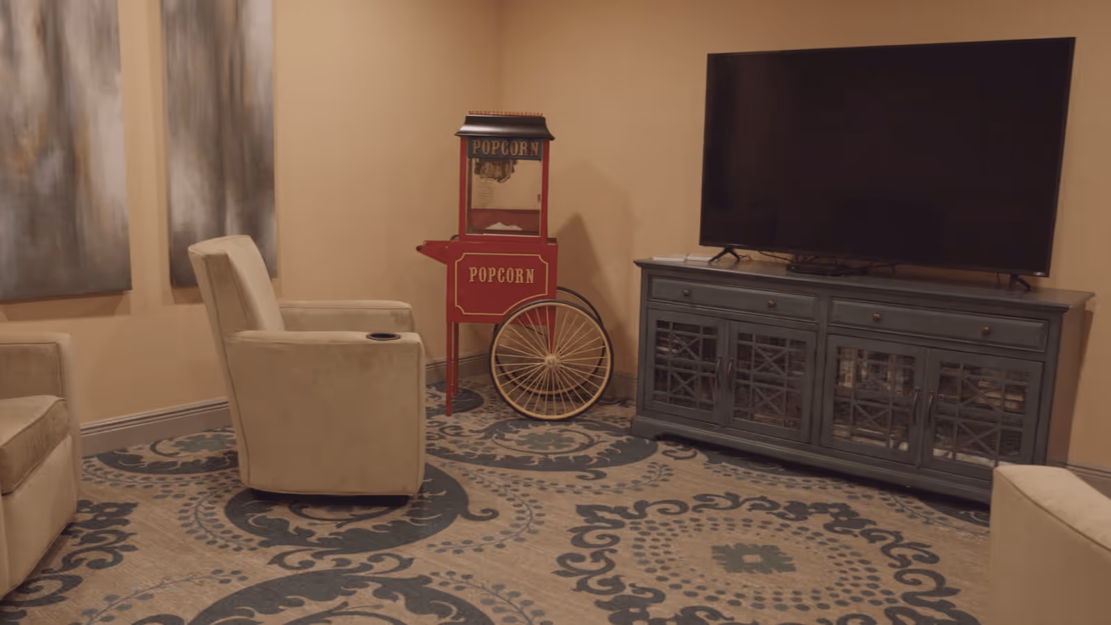 A cozy living room area with beige armchairs arranged around a patterned carpet. There is a large flat-screen TV on a dark wooden cabinet with glass-paneled doors. A vintage-style red popcorn machine with large wheels is placed against the wall. The walls are painted a warm beige color and there are two abstract paintings hanging on the left wall.