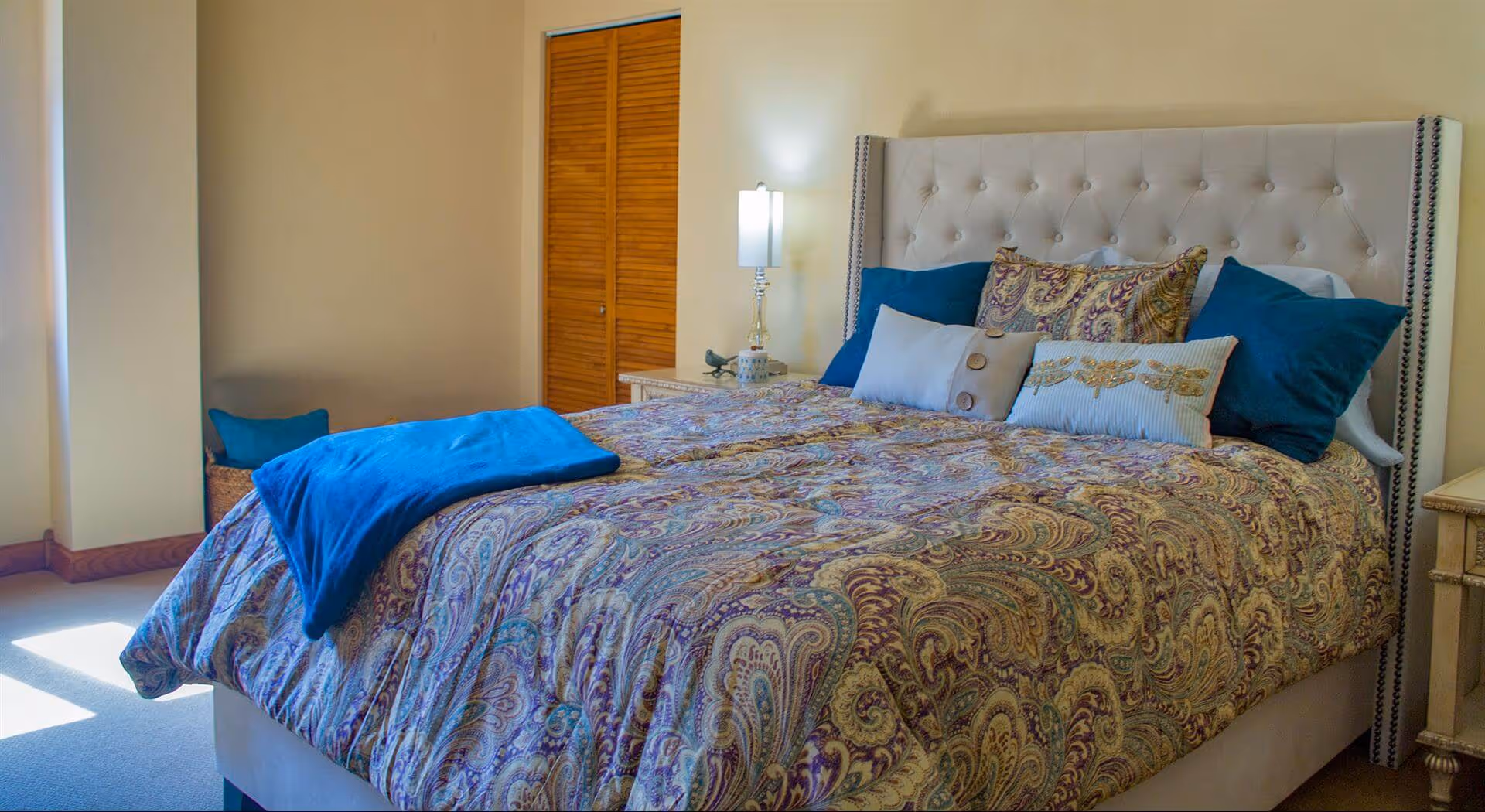 A bedroom with a large bed featuring a tufted white headboard. The bed is covered with a colorful paisley-patterned comforter and several decorative pillows in blue, beige, and gold tones. A blue throw blanket is draped over the foot of the bed. There is a nightstand with a lamp and decorative items next to the bed, and a wooden closet door is visible in the background. Natural light is coming in from a window on the left side.