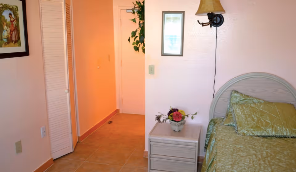 A bedroom corner in Azalea Manor featuring a bed with green quilted bedding and pillows, a small nightstand with a flower arrangement, a wall-mounted lamp, a framed picture, a closet with louvered doors, and a hallway leading to a closed door.