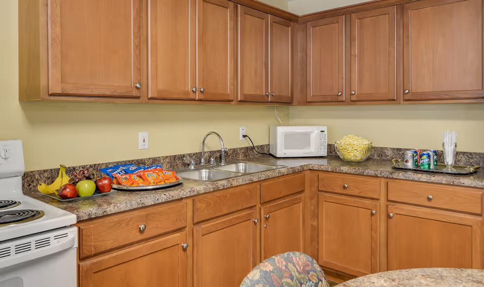 A kitchen area with wooden cabinets, a double sink, a white microwave, and a white electric stove. On the countertop, there are snacks including bags of chips, a bowl of popcorn, a plate with bananas, apples, and grapes, and a tray with soda cans and plastic utensils. A round table with a floral patterned chair is partially visible in the foreground.