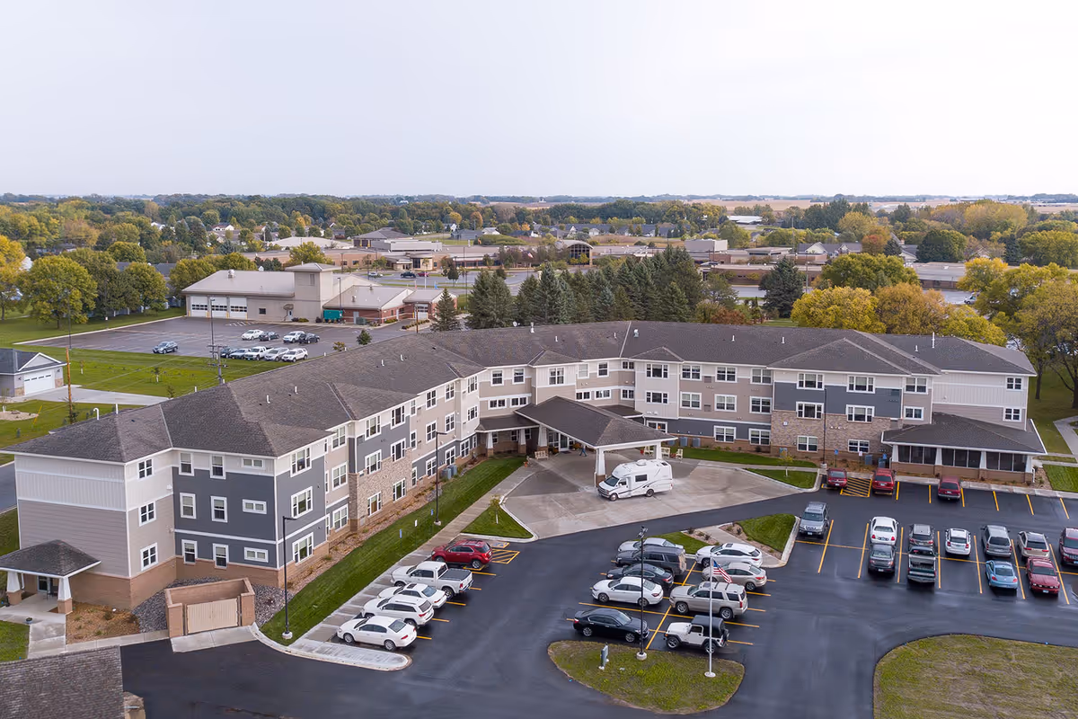 Aerial view of Brookside Senior Living, a large three-story senior living facility with a U-shaped building surrounded by parking lots filled with cars. The building has a covered entrance with a white van parked underneath. The surrounding area includes trees and other buildings in the distance under a cloudy sky.
