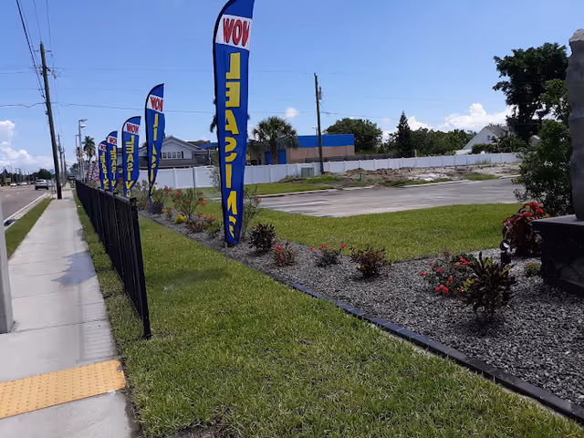Outdoor view of a grassy area with a sidewalk on the left and a row of tall blue and yellow flags that say 'WOW LEASE' along a black metal fence. There are small bushes and flowers planted in a gravel bed next to the grass. In the background, there is a parking lot and some buildings under a clear blue sky.