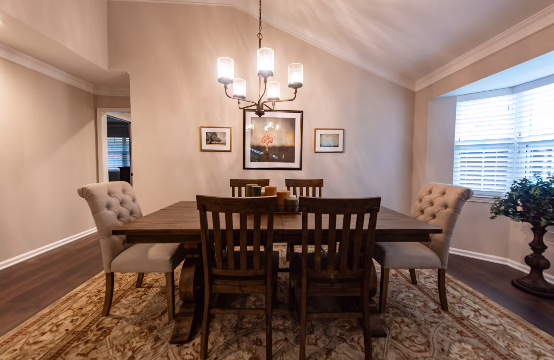 A dining room with a wooden table surrounded by six chairs, two upholstered and four wooden. A chandelier with five lights hangs above the table. The walls are beige with three framed pictures, and there is a large window with white blinds on the right side. A decorative plant stands in a pot near the window, and the floor is covered with a patterned rug.