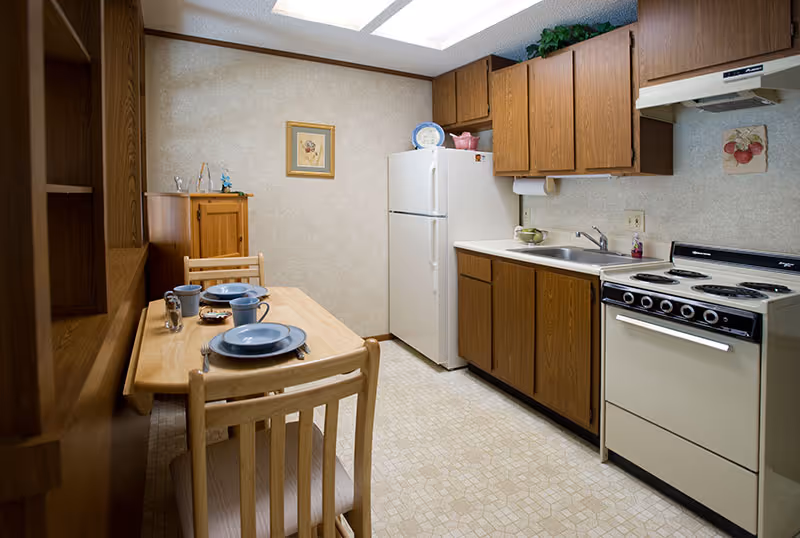 A small kitchen and dining area with wooden cabinets, a white refrigerator, a white stove with an oven, and a stainless steel sink. A wooden dining table with four chairs is set with blue plates, bowls, and mugs. The walls have light-colored wallpaper, and there is a framed picture hanging on the wall.