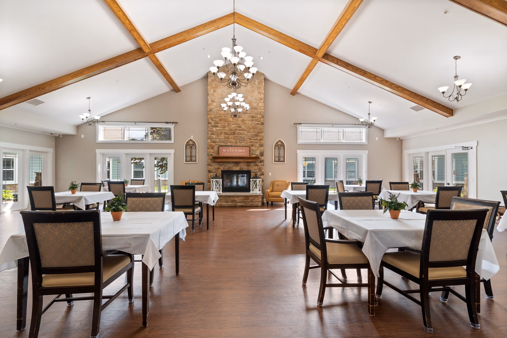 A spacious dining room with multiple tables covered in white tablecloths, each surrounded by chairs. The room features a high vaulted ceiling with wooden beams and several chandeliers. At the far end, there is a stone fireplace with a 'welcome' sign above it, flanked by decorative wall pieces. Large windows and glass doors allow natural light to fill the room, and small potted plants are placed on each table.
