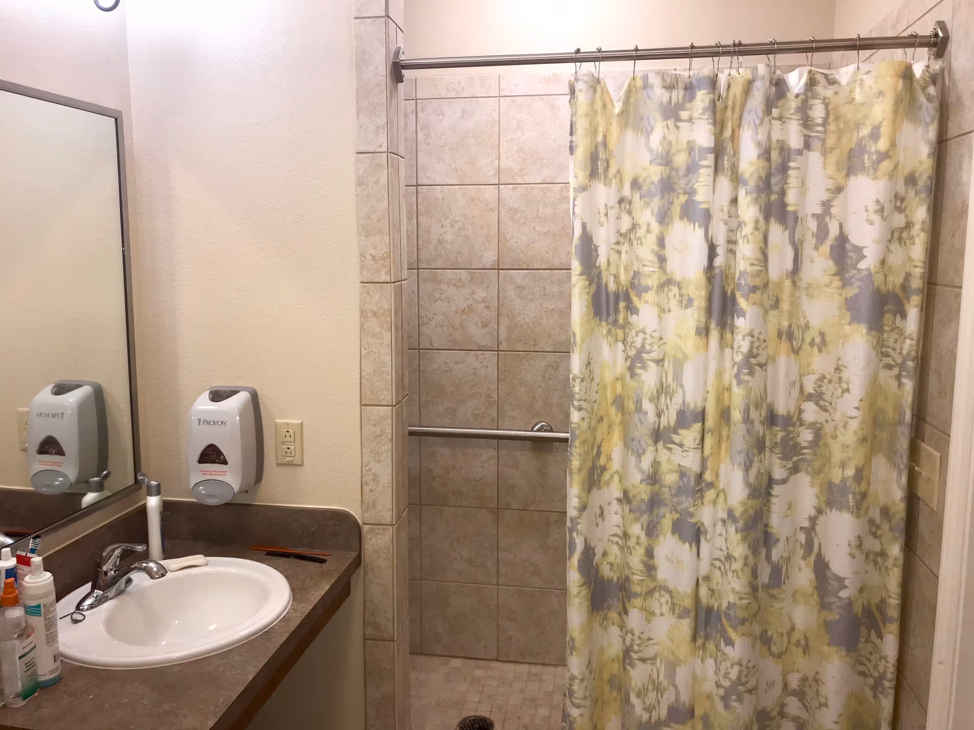 Small tiled bathroom with a sink and mirror on the left and a shower with a patterned curtain and grab bar on the right.