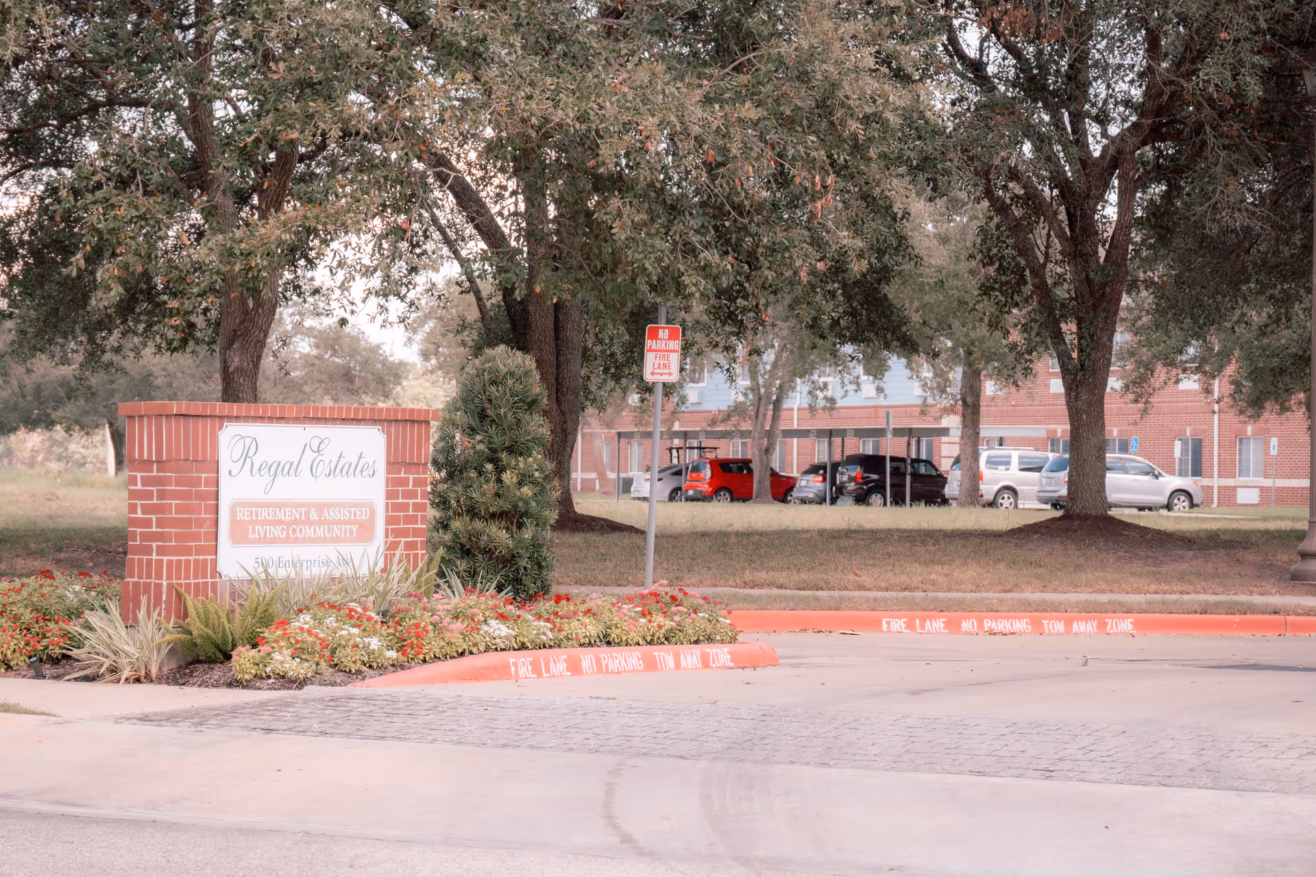 Entrance sign for Regal Estates Retirement & Assisted Living Community surrounded by flowers and greenery, with a parking area and trees in the background.