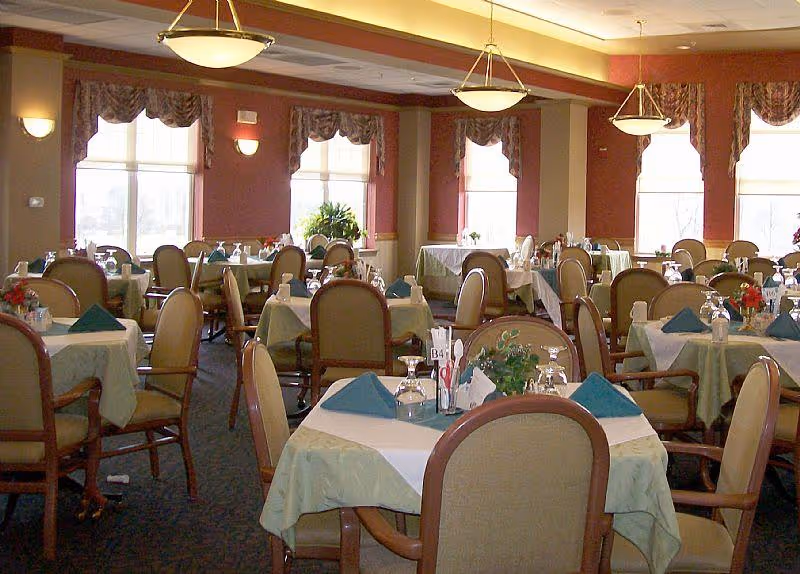 A dining room with multiple tables covered in light green tablecloths and white overlays, each set with blue folded napkins, glassware, and small floral centerpieces. The room has large windows with patterned valances, warm lighting fixtures on the ceiling and walls, and upholstered wooden chairs around the tables.