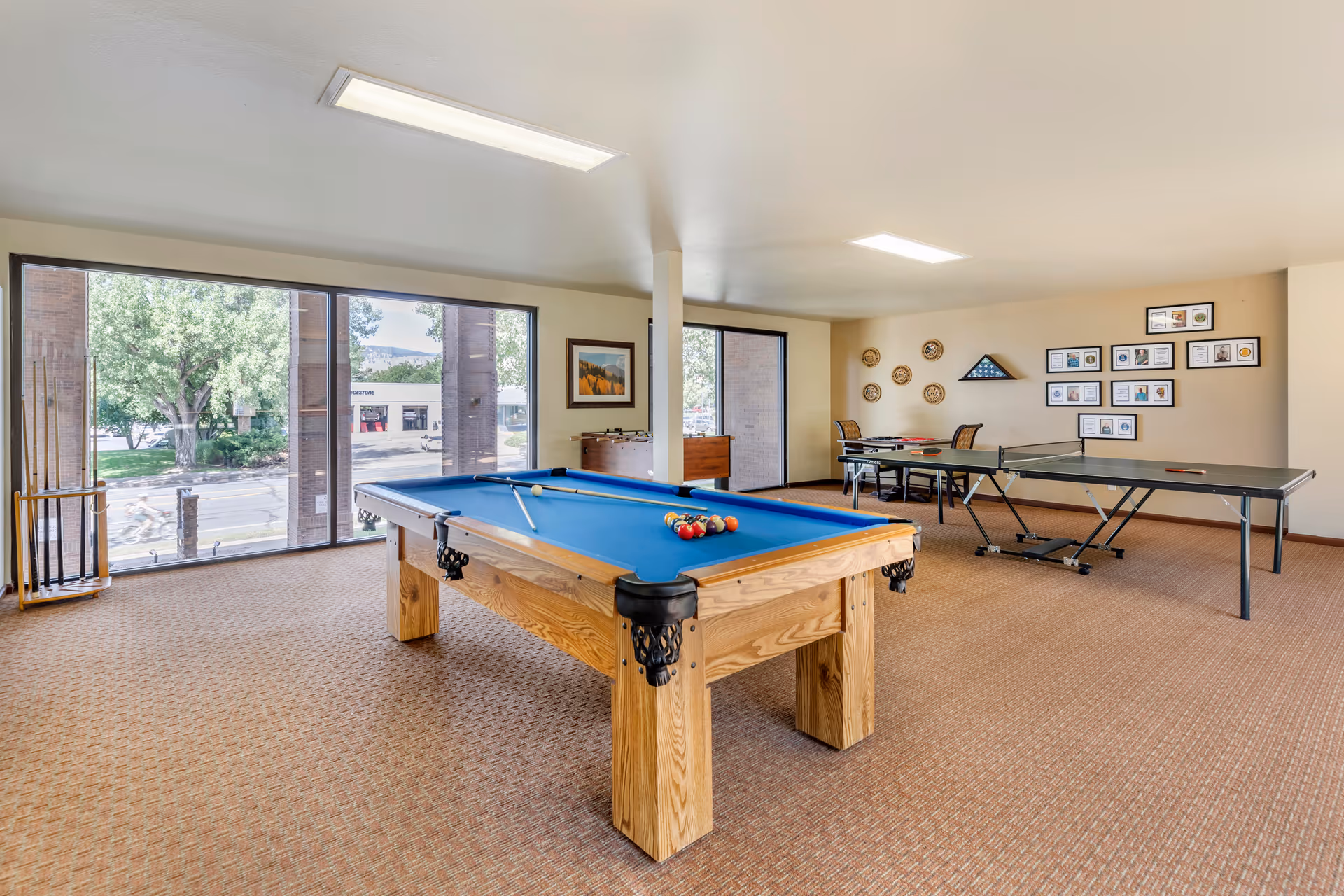 A bright recreation room with a pool table in the center, a ping pong table to the right, and a foosball table against the far wall. Large windows on the left side let in natural light and show trees and a street outside. The walls are decorated with framed pictures and a display of plaques and awards.