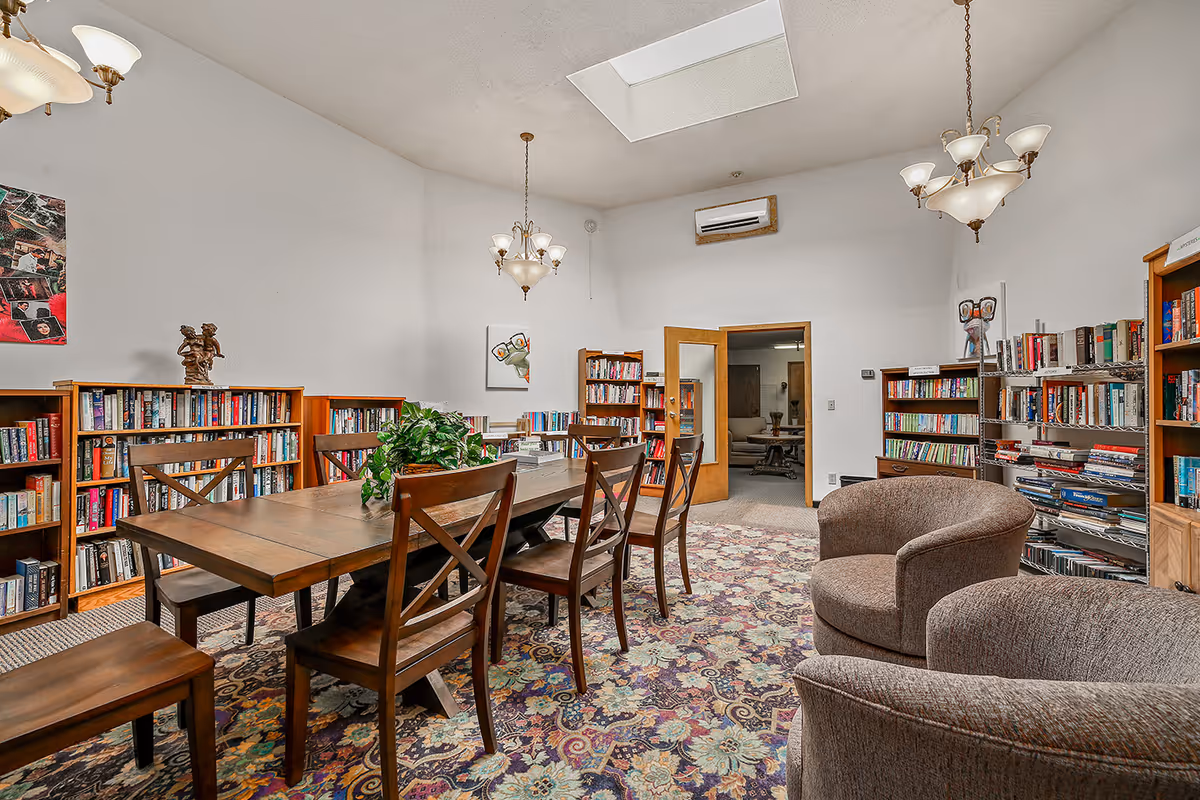 A bright communal library/reading room with bookshelves, a long wooden table and chairs, and upholstered armchairs under chandeliers and a skylight.