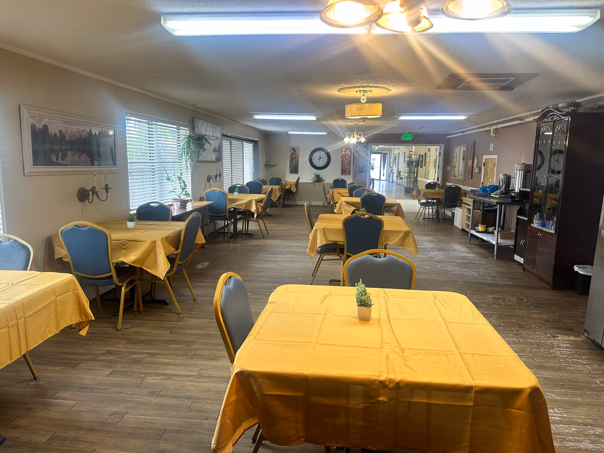 A dining room in an assisted living facility with multiple tables covered in yellow tablecloths and blue chairs around them. The room has wooden flooring, large windows with blinds on the left side, and various wall decorations including a large clock and framed pictures. There is a serving area with coffee dispensers and cups on the right side near the back of the room.