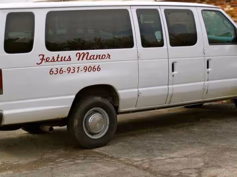 White Festus Manor van with red lettering and phone number parked outside.