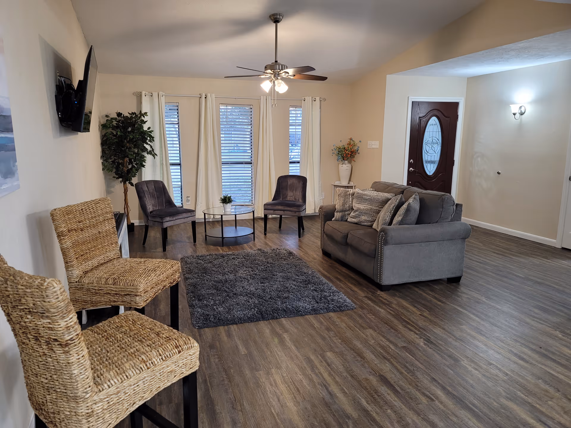 A cozy living room with a gray couch adorned with multiple pillows, two gray chairs, a round glass coffee table with a small plant, and two wicker chairs. The room has wood flooring, a ceiling fan with lights, three windows with white curtains, a dark wooden front door with an oval glass design, a potted plant, and a wall-mounted TV.