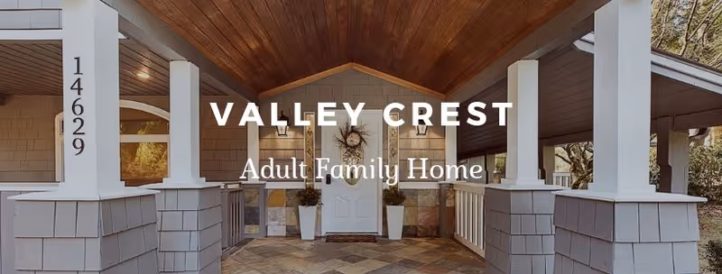 Front entrance of Valley Crest Adult Family Home featuring a covered porch with wooden ceiling, white pillars, stone accents, and a white door flanked by two potted plants.