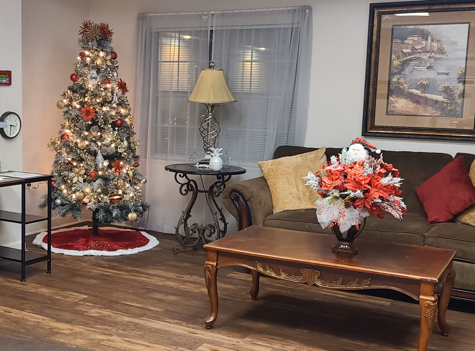 A cozy living room decorated for Christmas with a lit Christmas tree adorned with red and gold ornaments and a red tree skirt. There is a brown sofa with gold and red cushions, a wooden coffee table with a festive floral arrangement featuring a small Santa figure, a round side table with a lamp, and a framed painting on the wall. The floor is wooden and there is a window with sheer curtains behind the sofa.