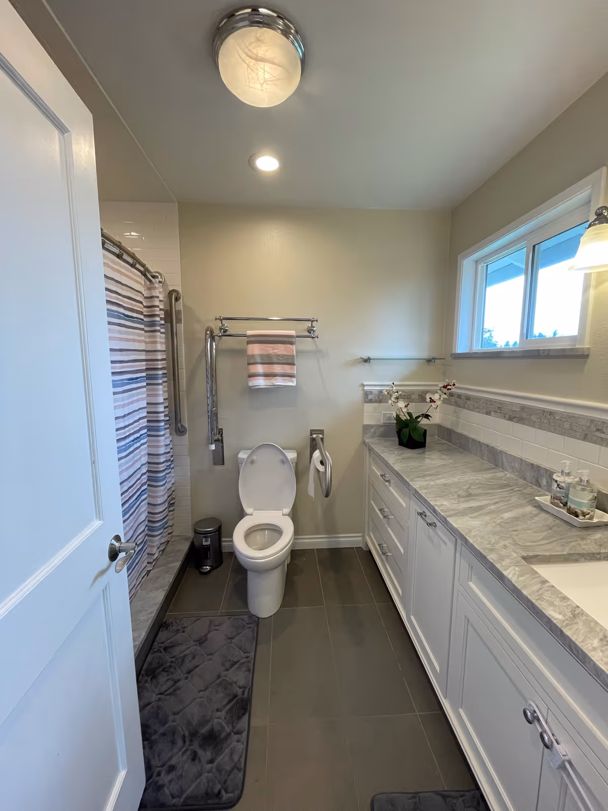 A clean and modern bathroom with a white toilet, a striped shower curtain, a towel rack with a striped towel, a gray floor mat, and a long marble countertop with white cabinets underneath. There is a window above the countertop and a small potted plant on the counter.