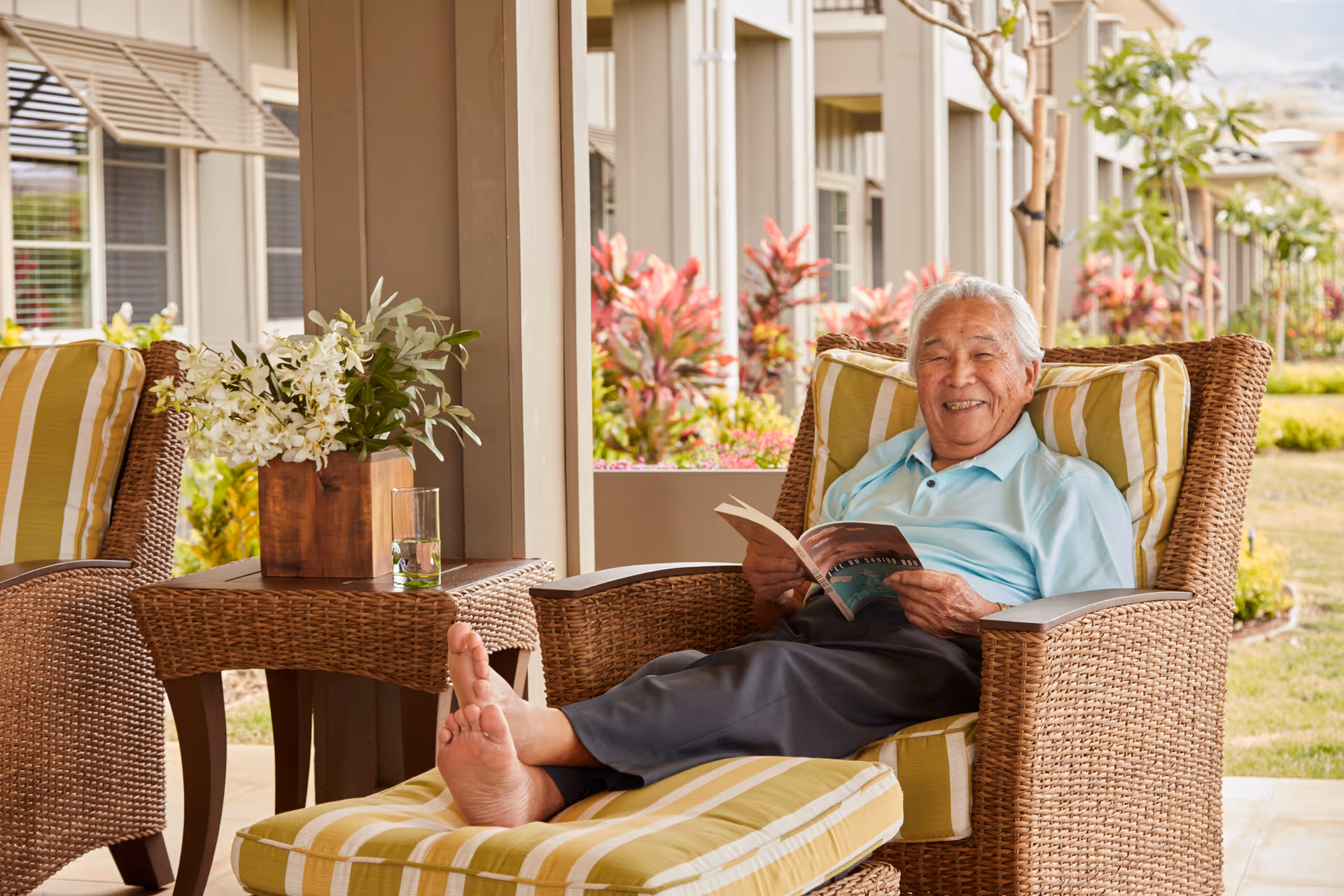An elderly man with gray hair is sitting barefoot on a cushioned wicker chair on a patio, smiling and holding an open book. Next to him is a matching wicker side table with a wooden vase of white flowers and a glass of water. The background shows a garden with colorful plants and the exterior of a building with windows.
