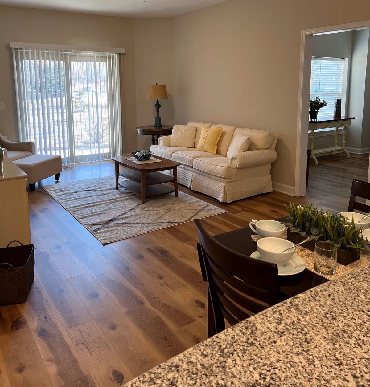 A cozy living room with a cream-colored sofa adorned with cushions, a wooden coffee table on a patterned rug, a side table with a lamp, and a sliding glass door with vertical blinds letting in natural light. Adjacent to the living room is a dining area with a granite countertop and place settings including bowls, plates, and glasses.