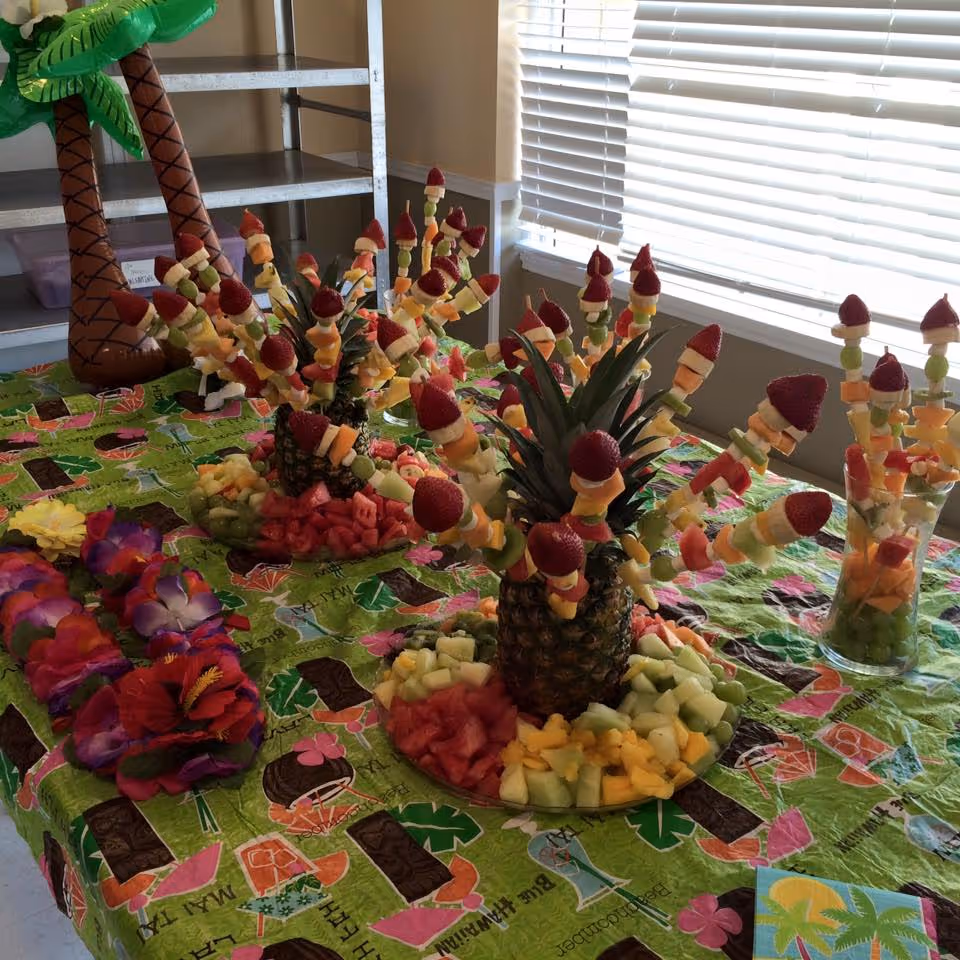 A decorated table with pineapple-centered fruit skewers, plates of cut fruit, leis and a tropical tablecloth in a bright room.