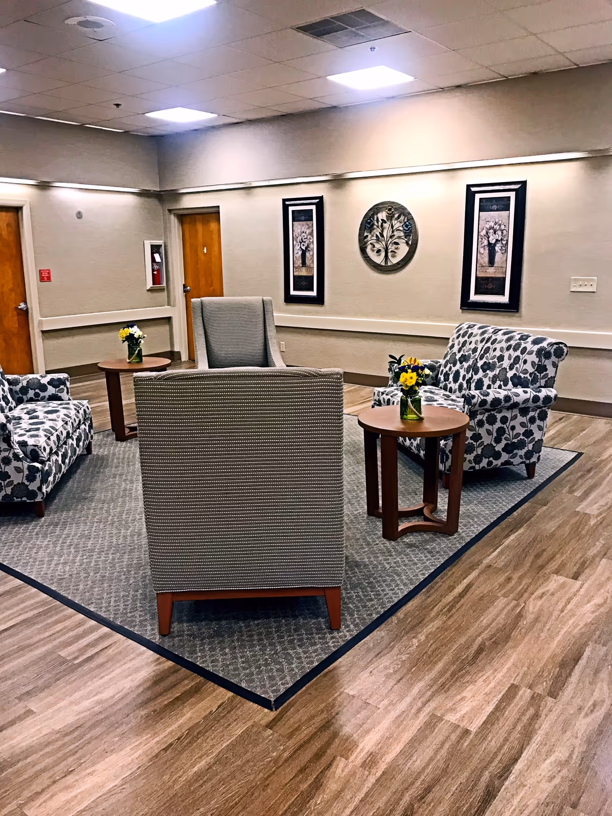 A cozy seating area in a senior living facility with four upholstered chairs arranged around two small wooden tables, each holding a small vase with yellow flowers. The room has light-colored walls adorned with three framed floral artworks and wood-look flooring with a large area rug under the seating. Two wooden doors are visible in the background.