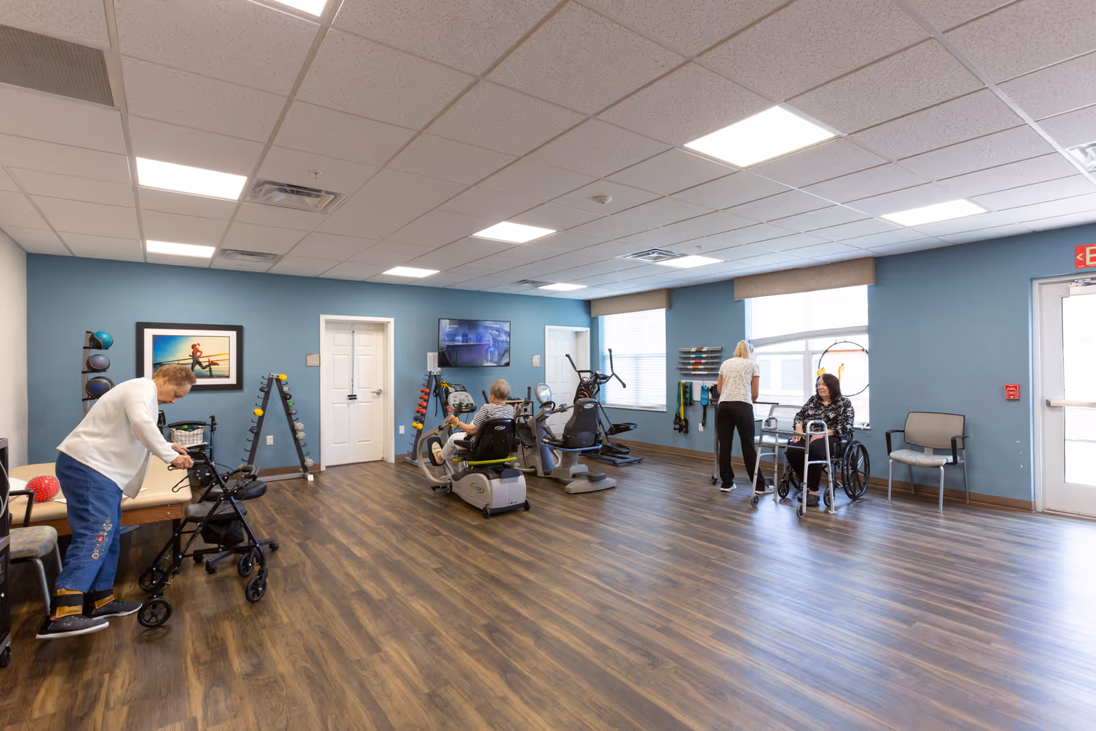 A spacious exercise room in a senior living facility with wood flooring and blue walls. The room contains exercise equipment including stationary bikes and weights. Four elderly individuals are present: one using a walker, one on a recumbent bike, one standing near the window, and one seated in a wheelchair. The room is well-lit with ceiling lights and natural light from windows.