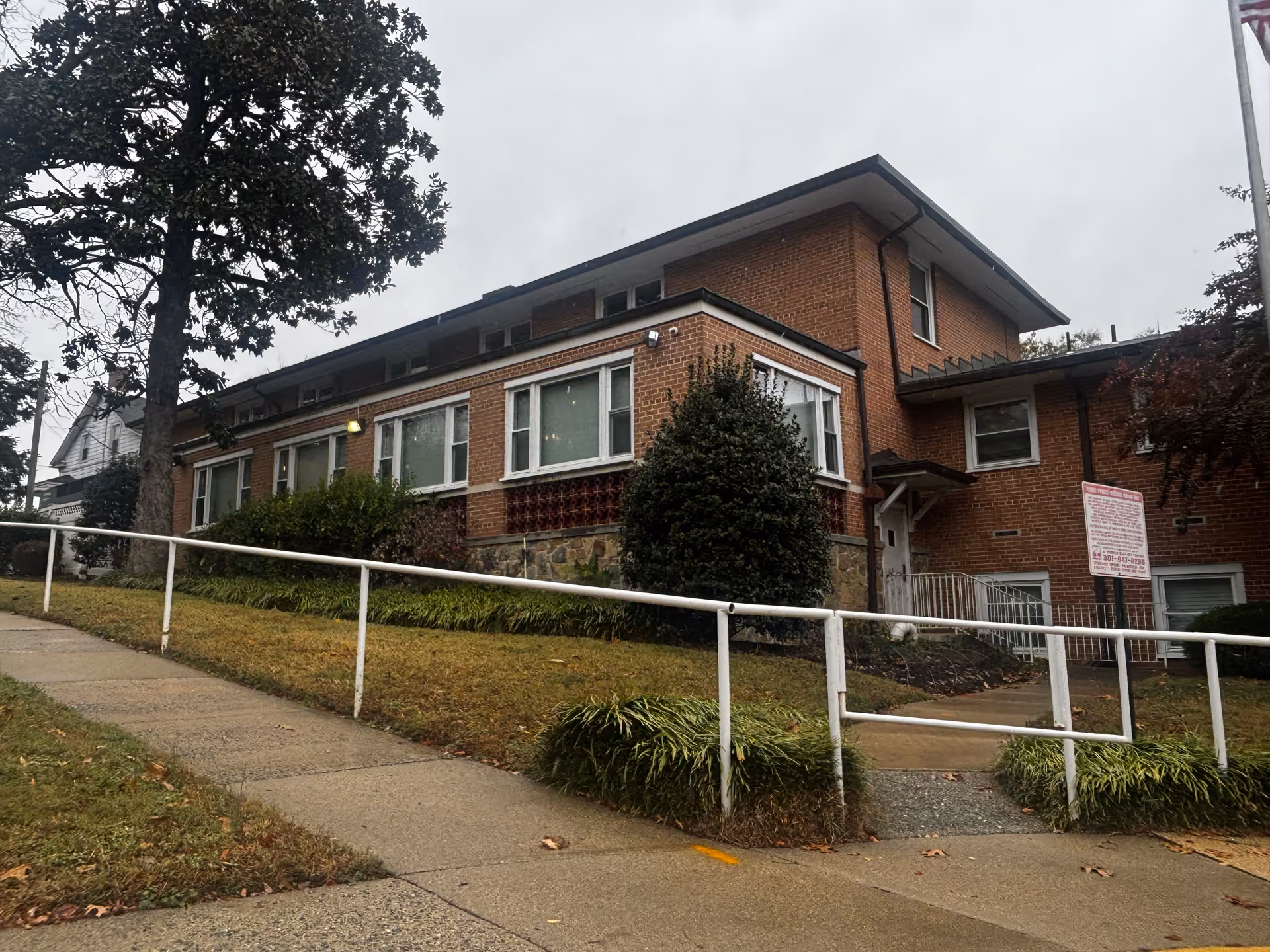 Exterior view of a two-story brick building with multiple windows, surrounded by bushes and a tree. A concrete sidewalk with a white metal handrail runs alongside the building on a slight incline. The sky is overcast.