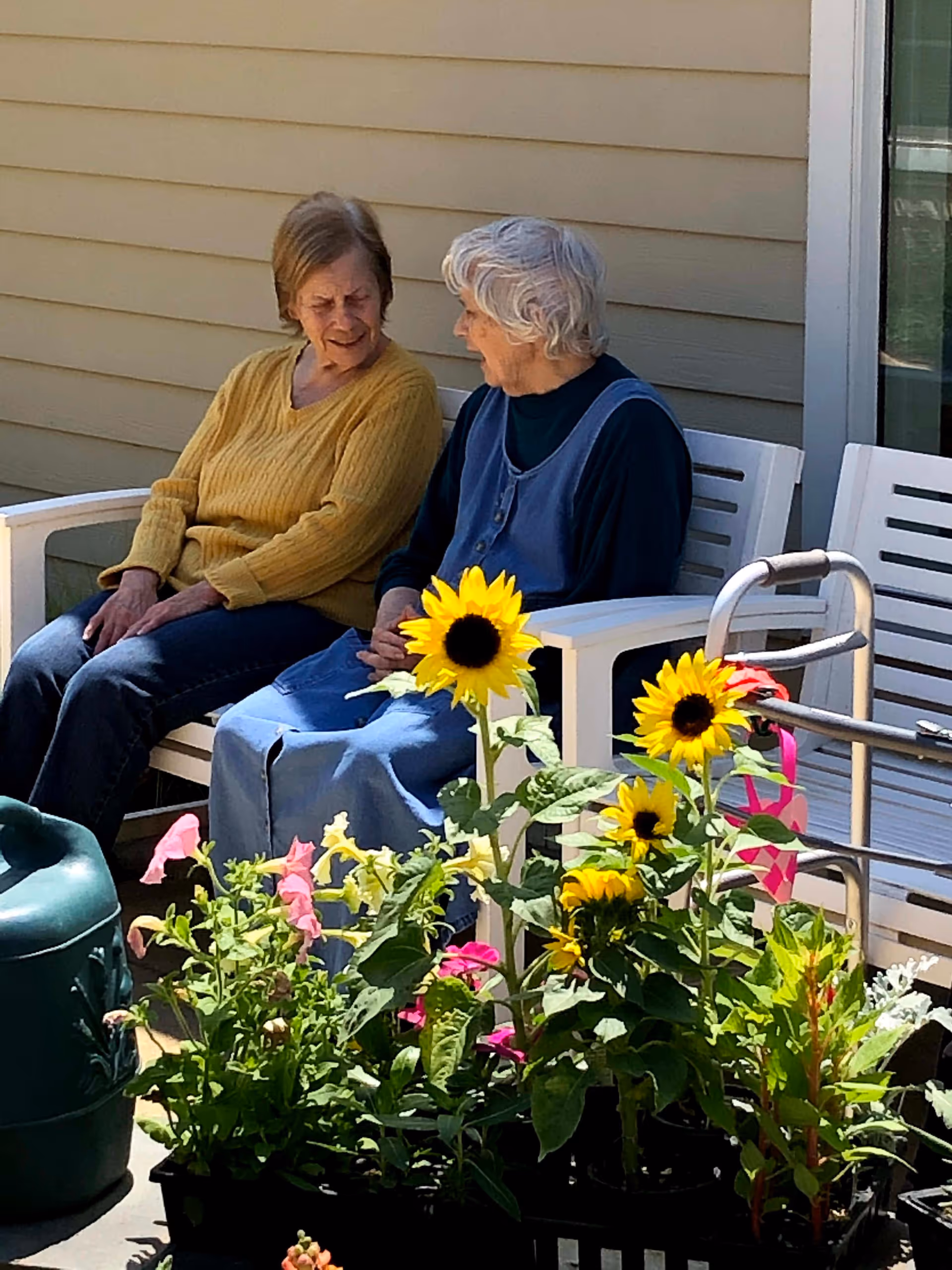Two elderly women sitting on a white bench outside a building, engaged in conversation. In front of them are various potted flowers including sunflowers and pink blooms. A walker is positioned next to the bench.
