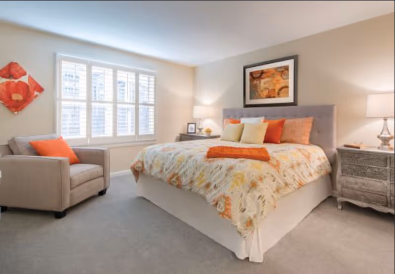 Bright, well-decorated bedroom with a large bed, bedside tables and lamps, an armchair, and a window with shutters.
