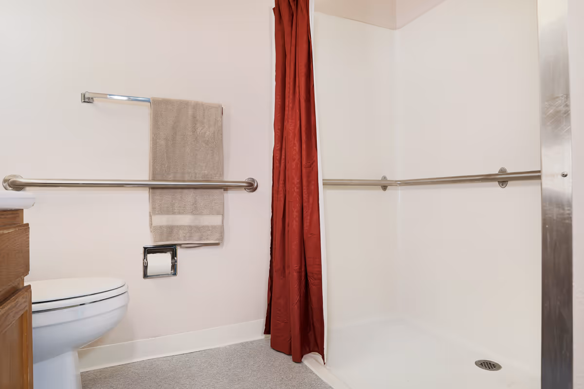 Accessible bathroom with a toilet, horizontal grab bars, a towel on a rack, and a shower with a red curtain.