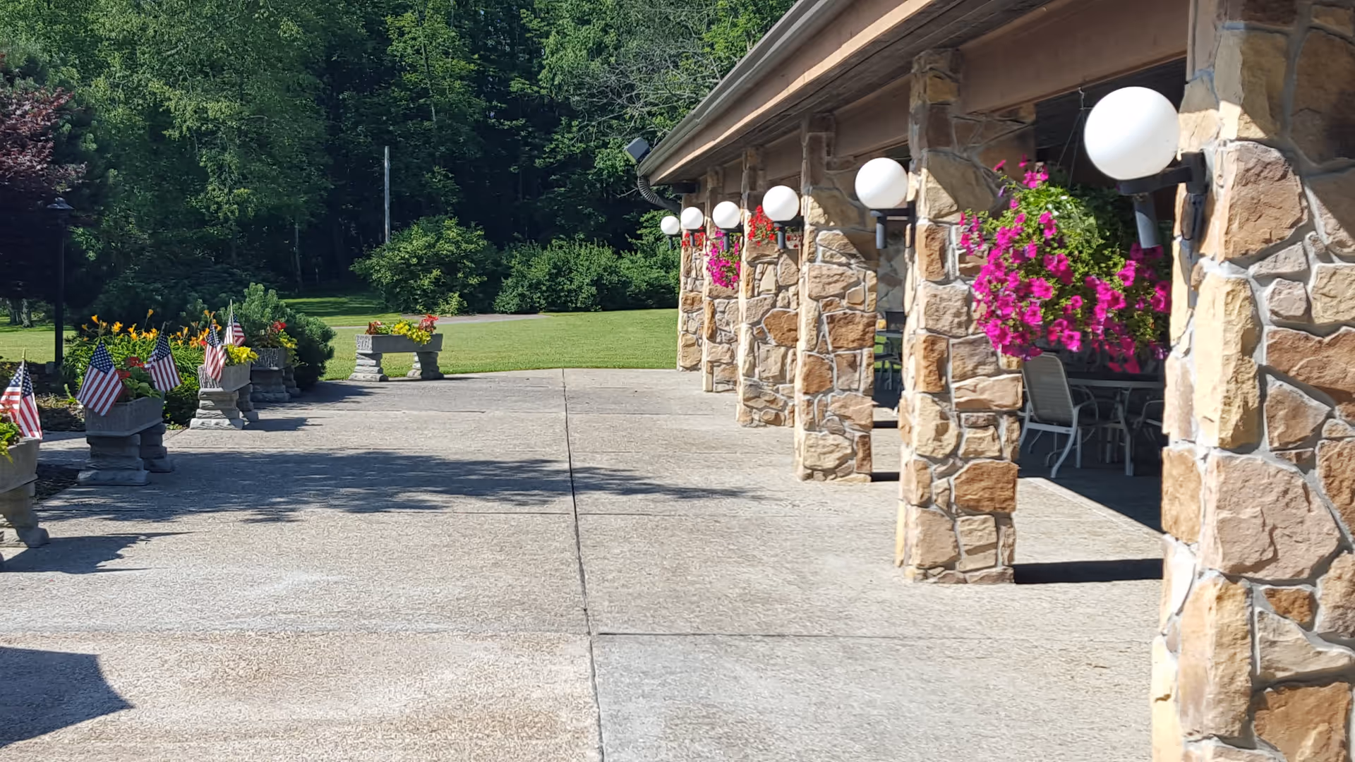 Stone-pillared covered patio with hanging flower baskets, outdoor seating and planters with American flags beside a lawn.