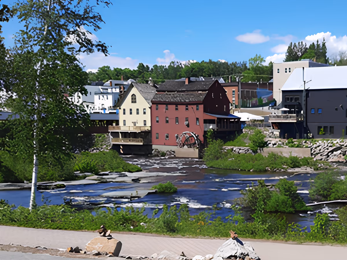 View of a river with rocks and greenery along the banks, with several buildings in the background including a prominent red building with a water wheel. Trees and a clear blue sky with some clouds are visible.