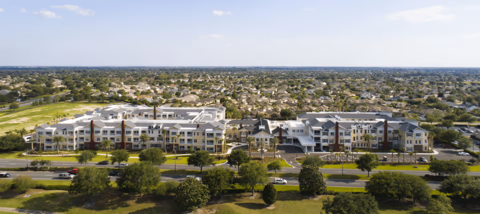 Aerial view of a large senior living facility named Watercrest Buena Vista, featuring multiple connected buildings with white and beige exteriors, surrounded by trees, parking areas, and a suburban neighborhood in the background under a clear blue sky.