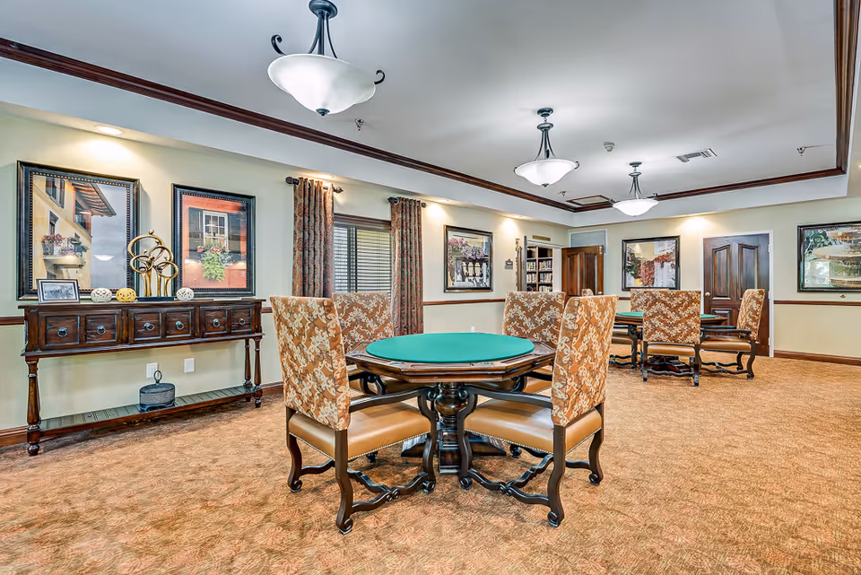 Well-lit common room with round green-felt game tables, upholstered chairs, framed artwork and a decorative sideboard.