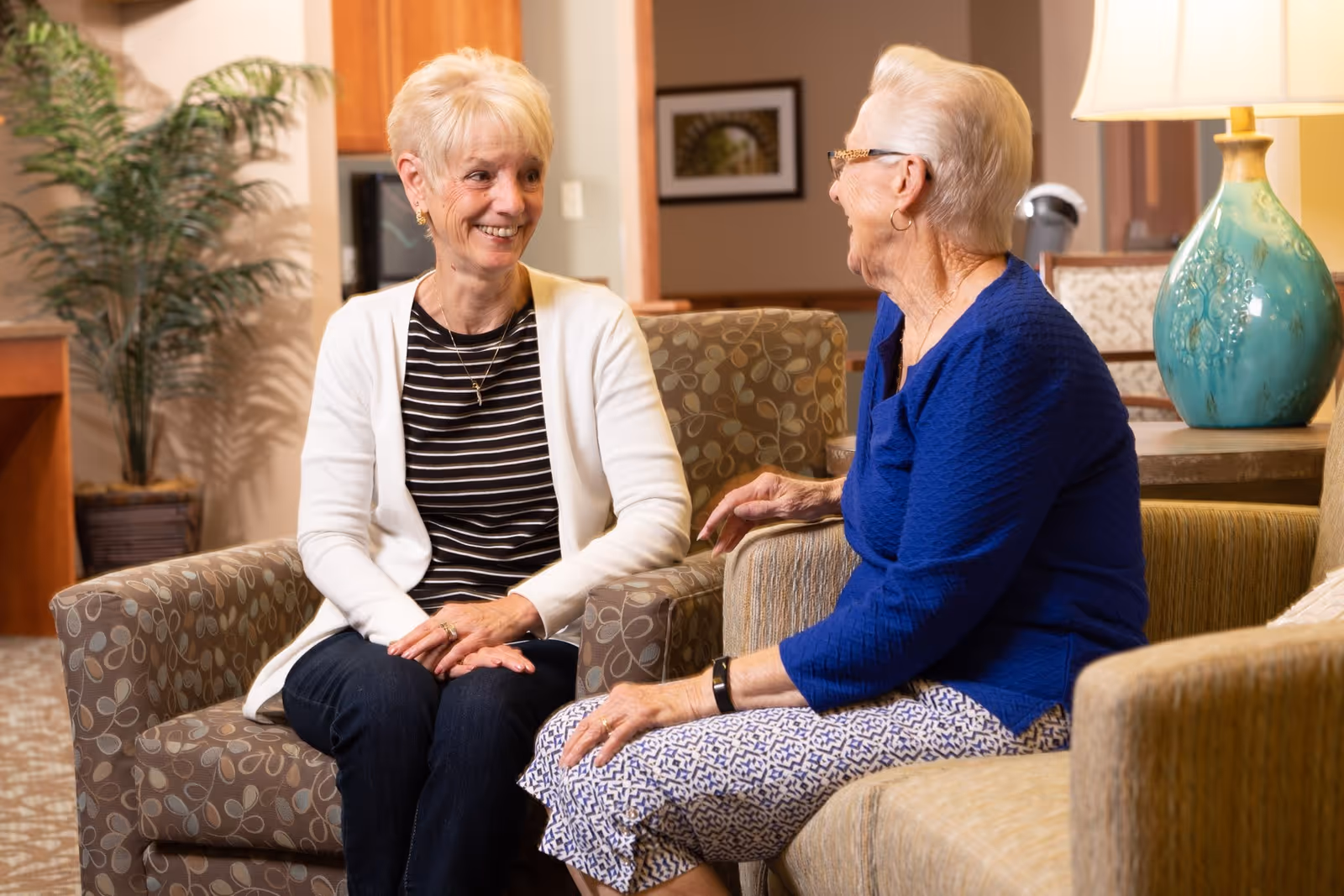 Two elderly women sitting and talking in a cozy living room area with patterned armchairs, a lamp on a side table, and a potted plant in the background.
