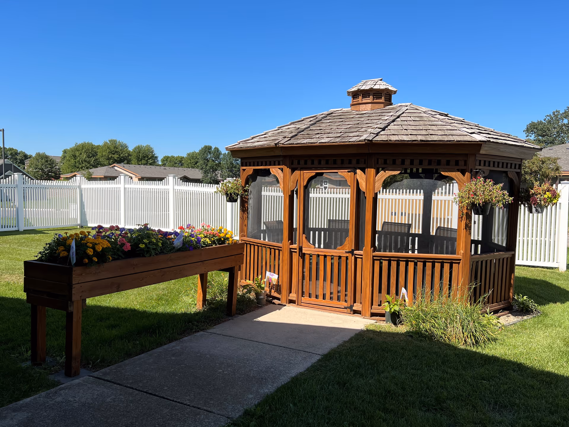 A wooden gazebo with a shingled roof and screened windows sits on a concrete path in a grassy yard. There is a raised wooden planter box filled with colorful flowers to the left of the gazebo. The area is enclosed by a white picket fence, and the sky is clear and blue.
