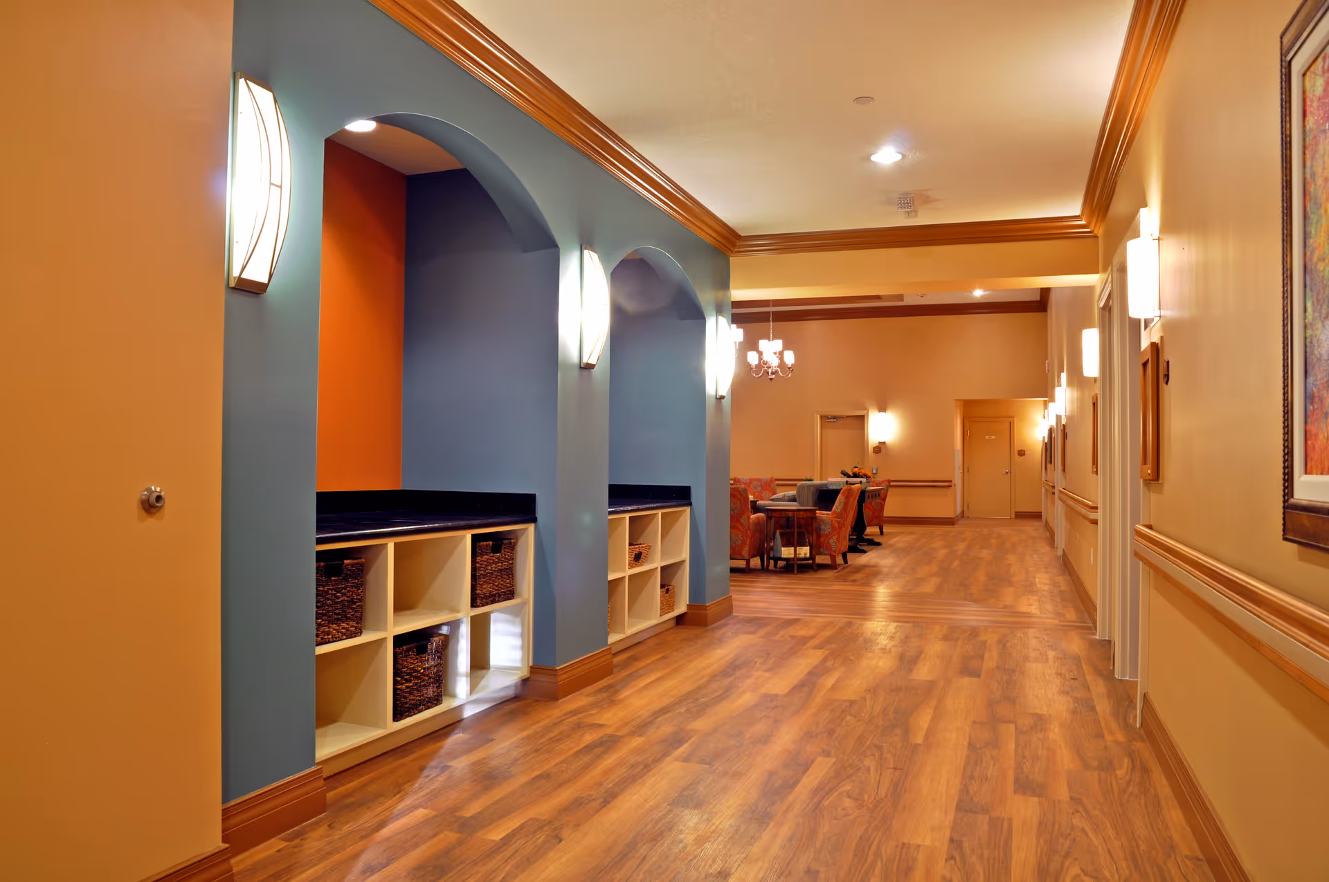 A well-lit hallway in a senior living facility with wooden flooring, beige and blue walls, and decorative wall sconces. There are built-in shelves with baskets on the left side under blue archways, and a seating area with chairs and a table is visible at the end of the hallway.