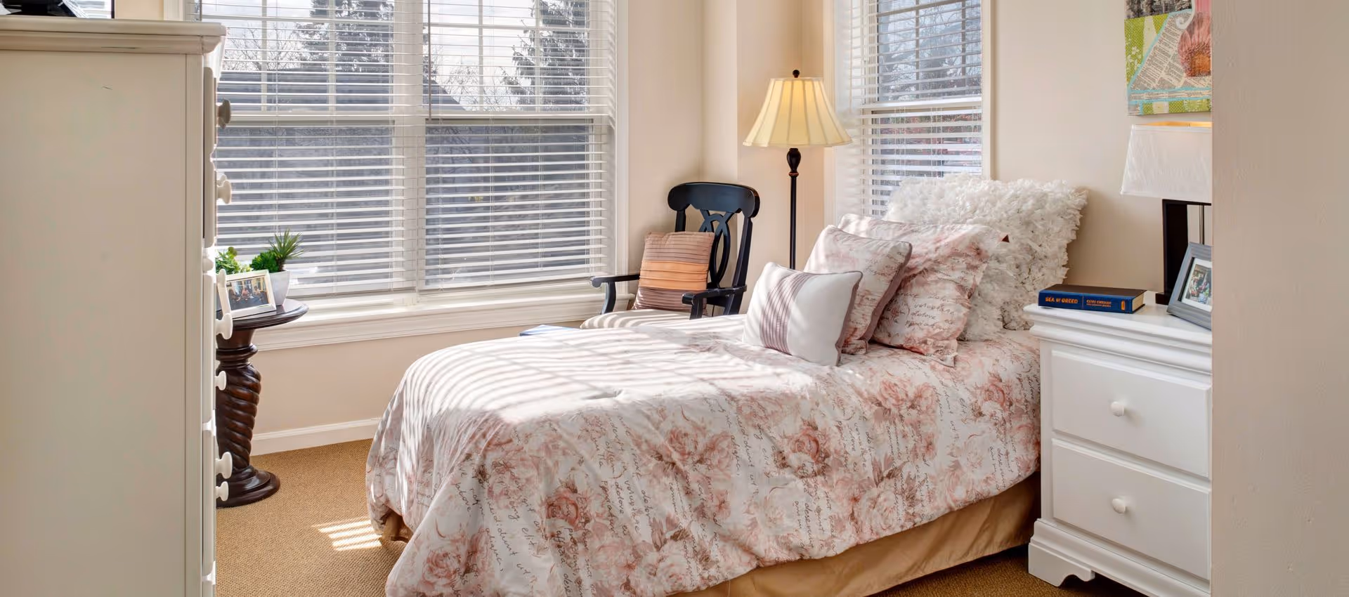 Sunlit cozy bedroom with a single bed dressed in floral bedding, a bedside table and lamp, a chair, and large windows with blinds.