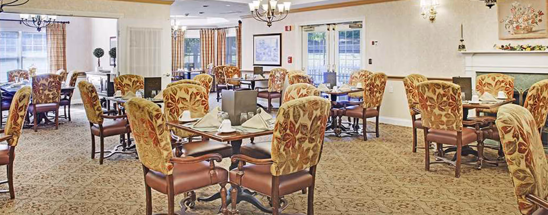 A spacious dining room with multiple tables set for meals, each table surrounded by upholstered chairs with floral patterns. The room has carpeted floors, chandeliers, large windows with curtains, and a fireplace on the right side.