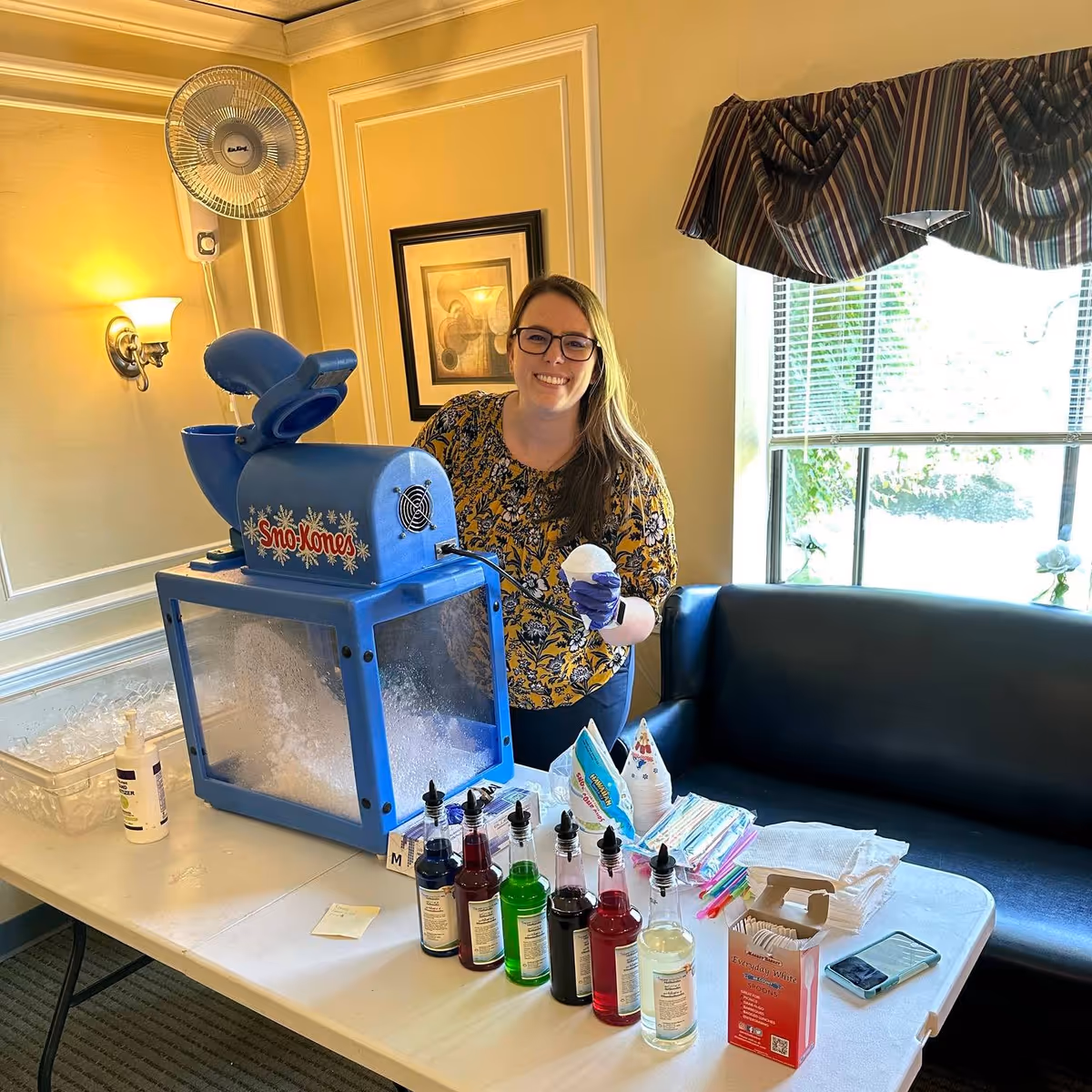 A woman wearing glasses and a yellow floral blouse stands behind a table with a blue Sno-Konas shaved ice machine. She is holding a cup of shaved ice and smiling. The table also has various bottles of flavored syrups, napkins, straws, and a hand sanitizer bottle. The setting appears to be a cozy indoor room with a window, a black couch, and warm lighting.