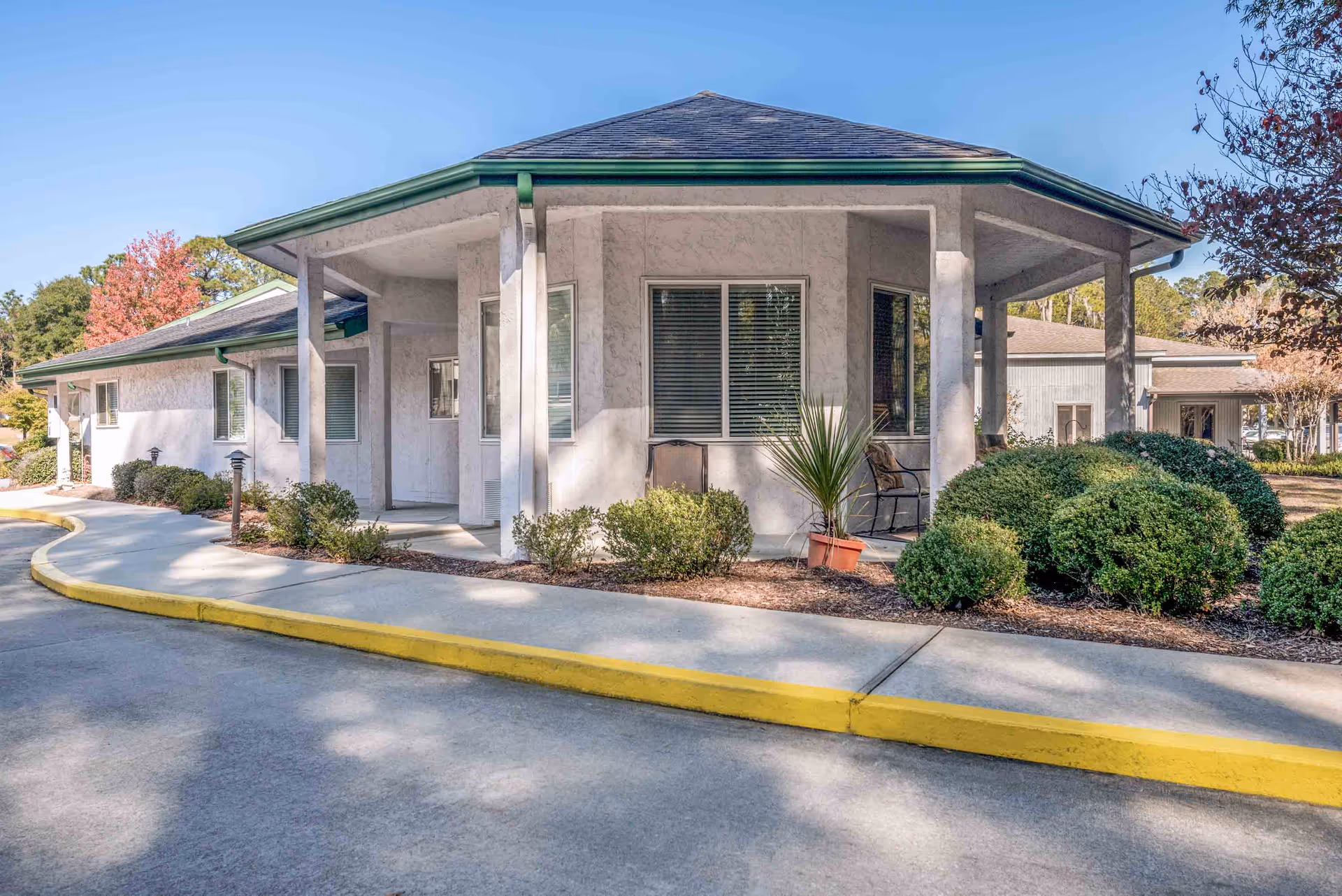 Exterior view of a single-story building with a covered porch area, white walls, green trim, and a gray roof. The building is surrounded by neatly trimmed bushes and small trees, with a curved sidewalk and a yellow-painted curb in front.