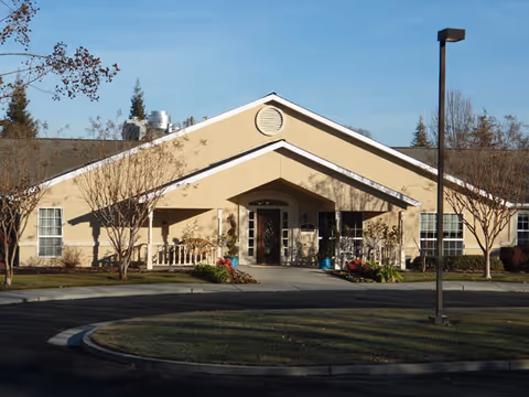 Exterior front view of a single-story beige building with a peaked roof and an entrance covered by a small porch. There are leafless trees and shrubs around the building, a curved driveway in front, and a tall street lamp on the right side. The sky is clear and blue.