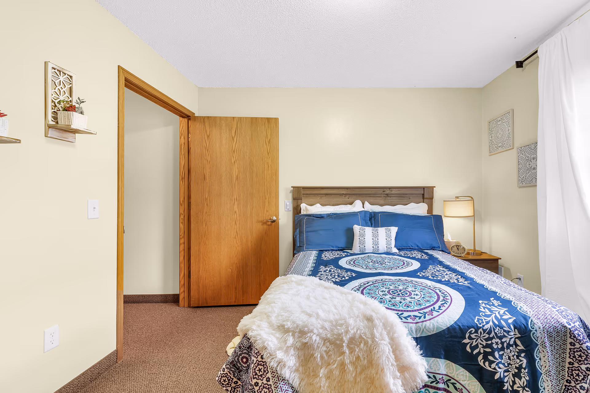 A cozy bedroom with a bed covered in a blue patterned comforter and white pillows. There is a wooden headboard, a nightstand with a lamp and clock, beige walls with decorative wall art, a wooden door slightly open, and a window with white curtains letting in natural light.