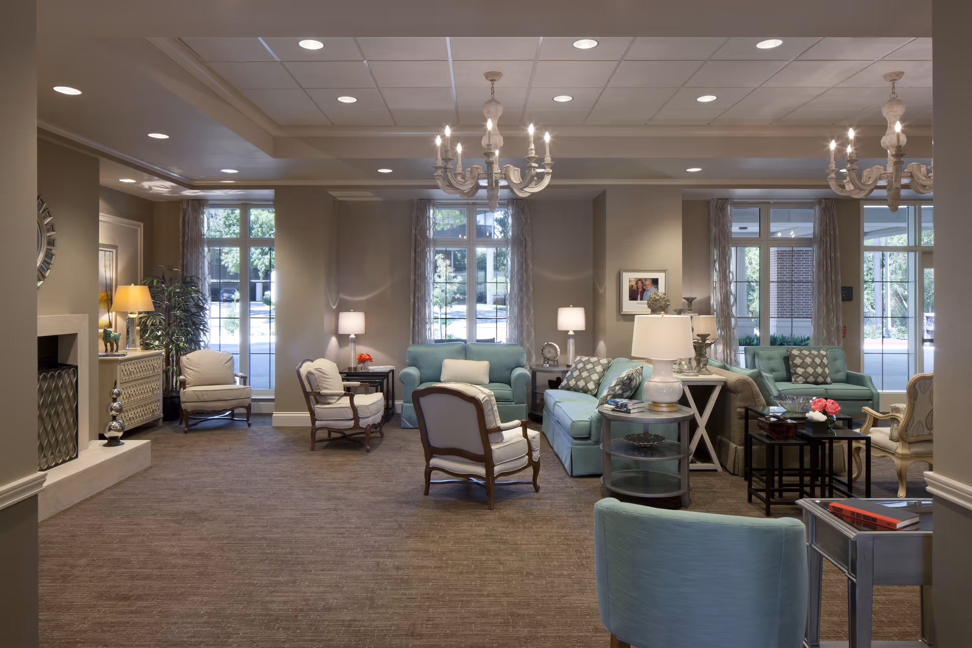 Spacious senior living common room with teal sofas and armchairs arranged around side tables, chandeliers, lamps, and large windows.