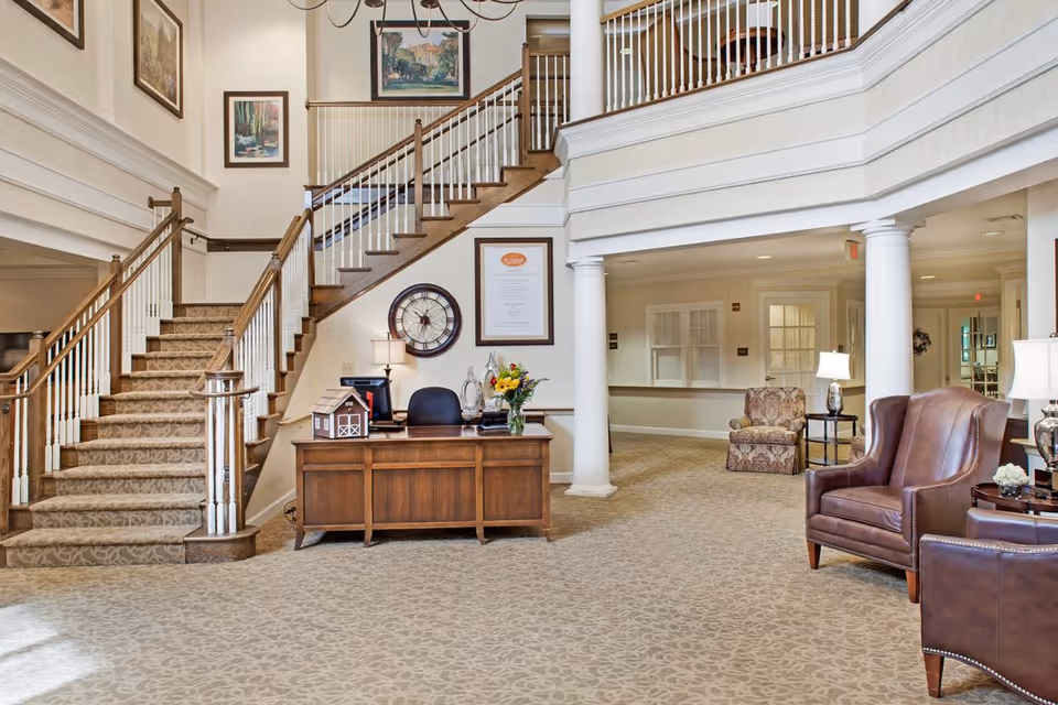 A spacious and well-lit interior lobby area featuring a wooden reception desk with a computer, a vase of flowers, and a decorative miniature barn. Behind the desk is a large wall clock and framed artwork. To the left, a carpeted staircase with wooden handrails leads to an upper floor. The area includes comfortable leather and fabric armchairs, side tables with lamps, and white columns supporting the upper balcony. The walls are adorned with framed paintings.